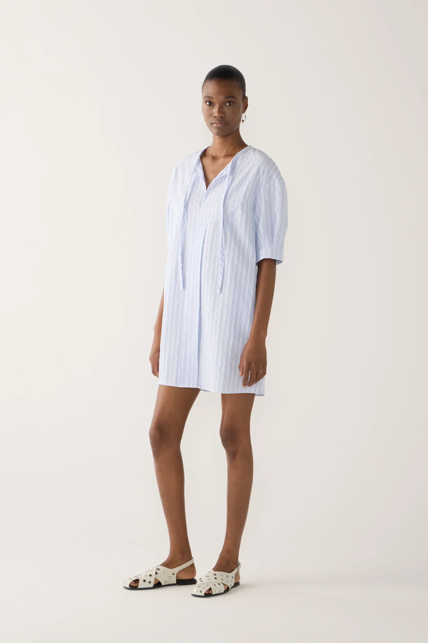 Side view of model wearing mini dress in blue and white striped seersucker fabric with drop shoulder, cap sleeve detail, front tie, side seam pockets.