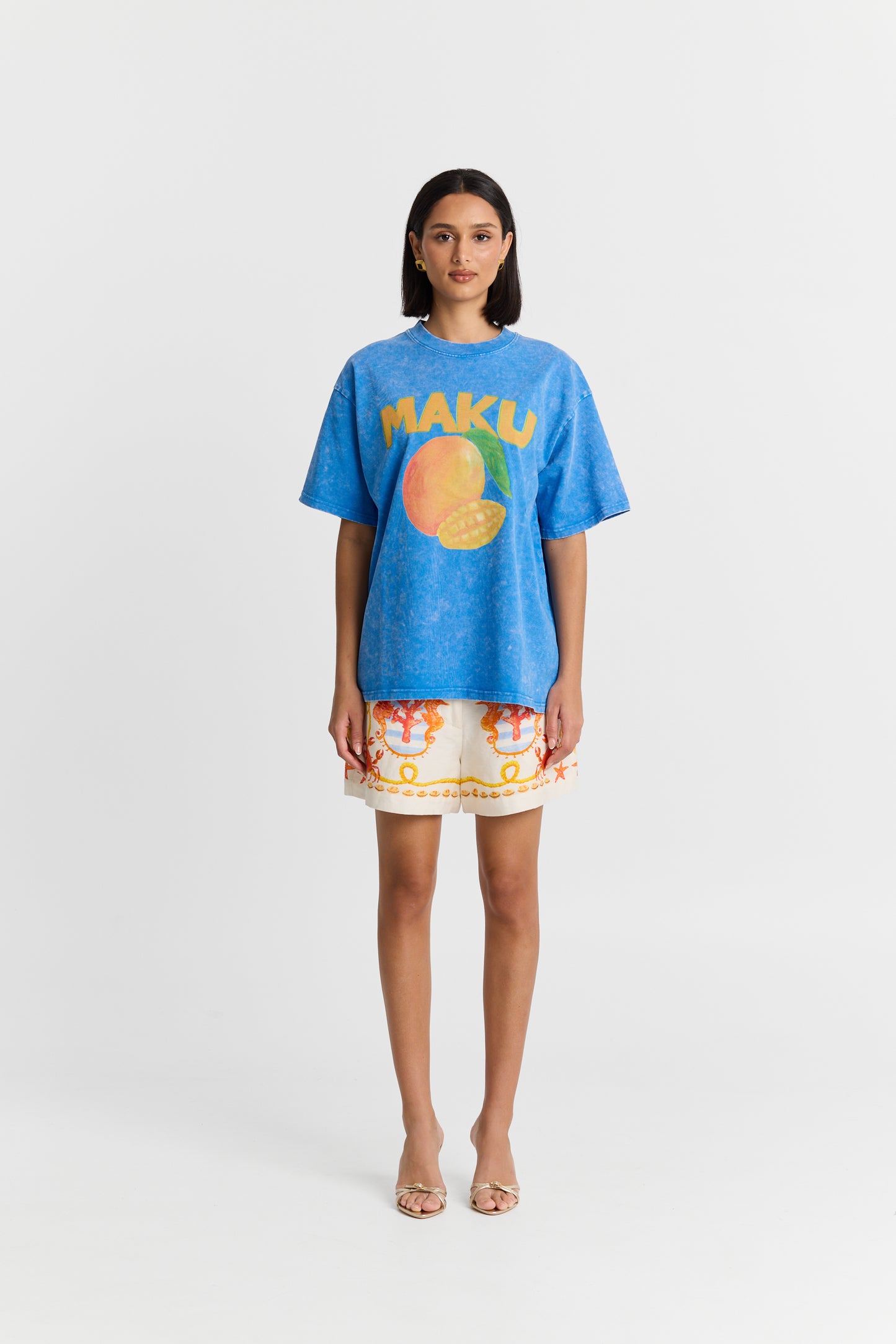 A Woman wearing a blue t-shirt with 'MAKU' and a mango graphic, paired with colorful shorts on a white background