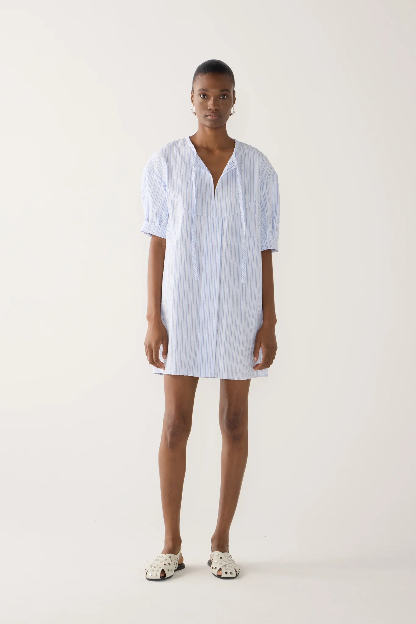 Model wearing mini dress in  blue and white striped seersucker fabric with drop shoulder, cap sleeve detail, front tie, side seam pockets.