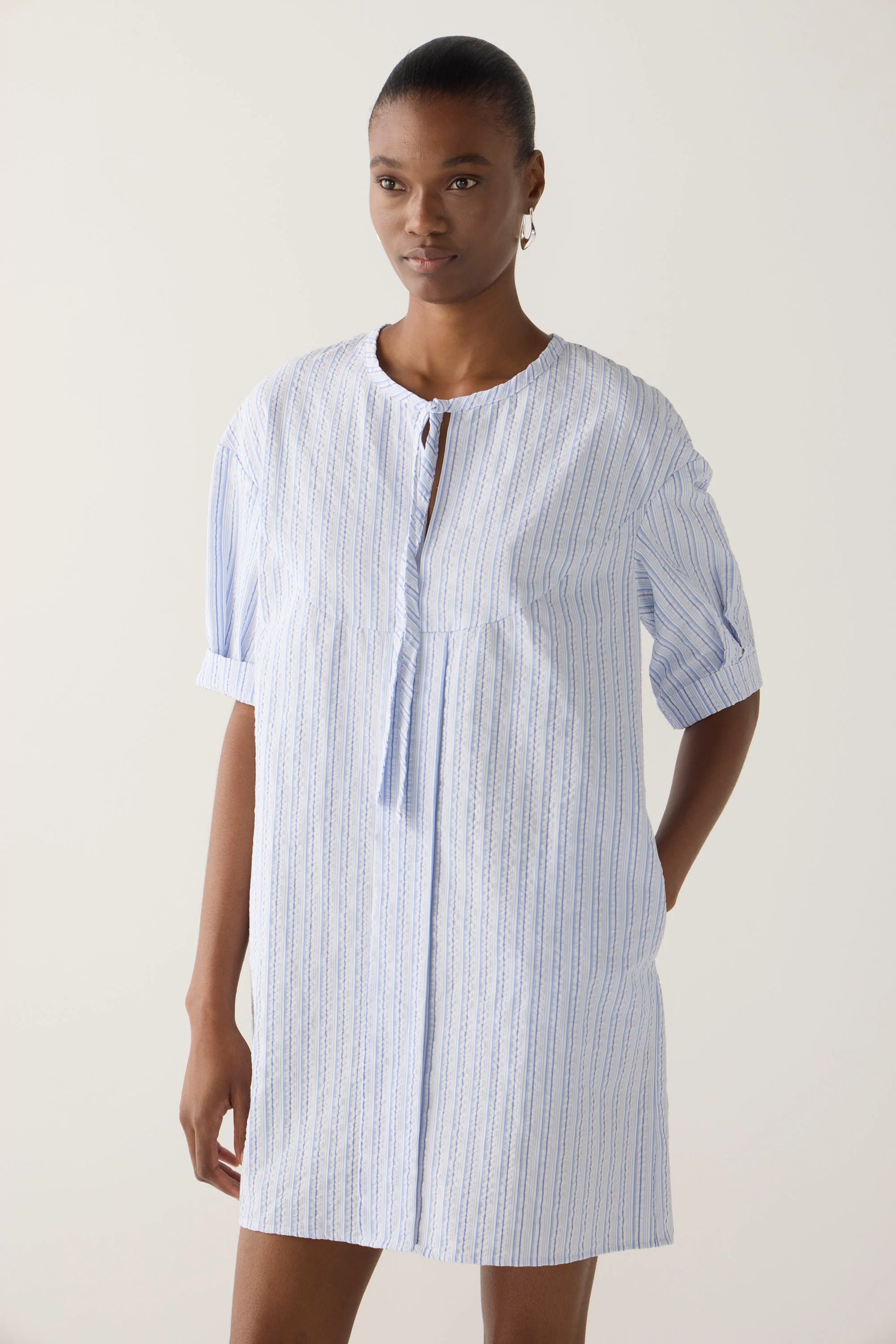 Close up of model wearing mini dress in  blue and white striped seersucker fabric with drop shoulder, cap sleeve detail, front tie, side seam pockets.