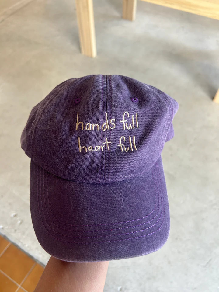 Purple cap with white “hands full, heart full” text made from cotton