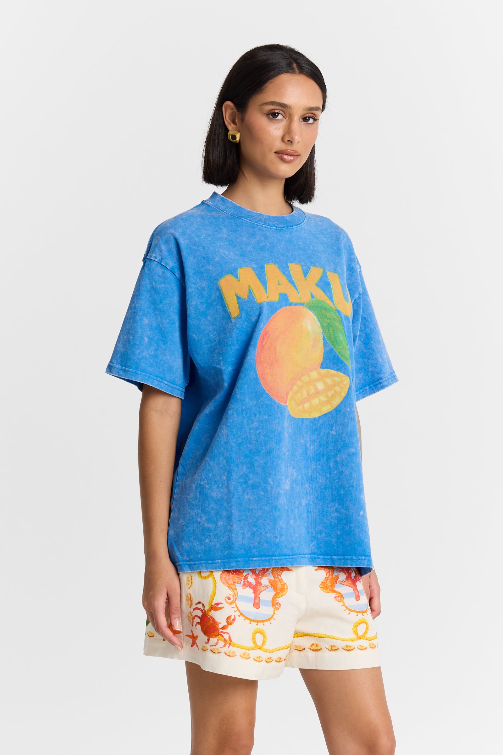 A Woman wearing a blue t-shirt with 'MAKU' and a mango graphic, paired with colorful shorts on a white background