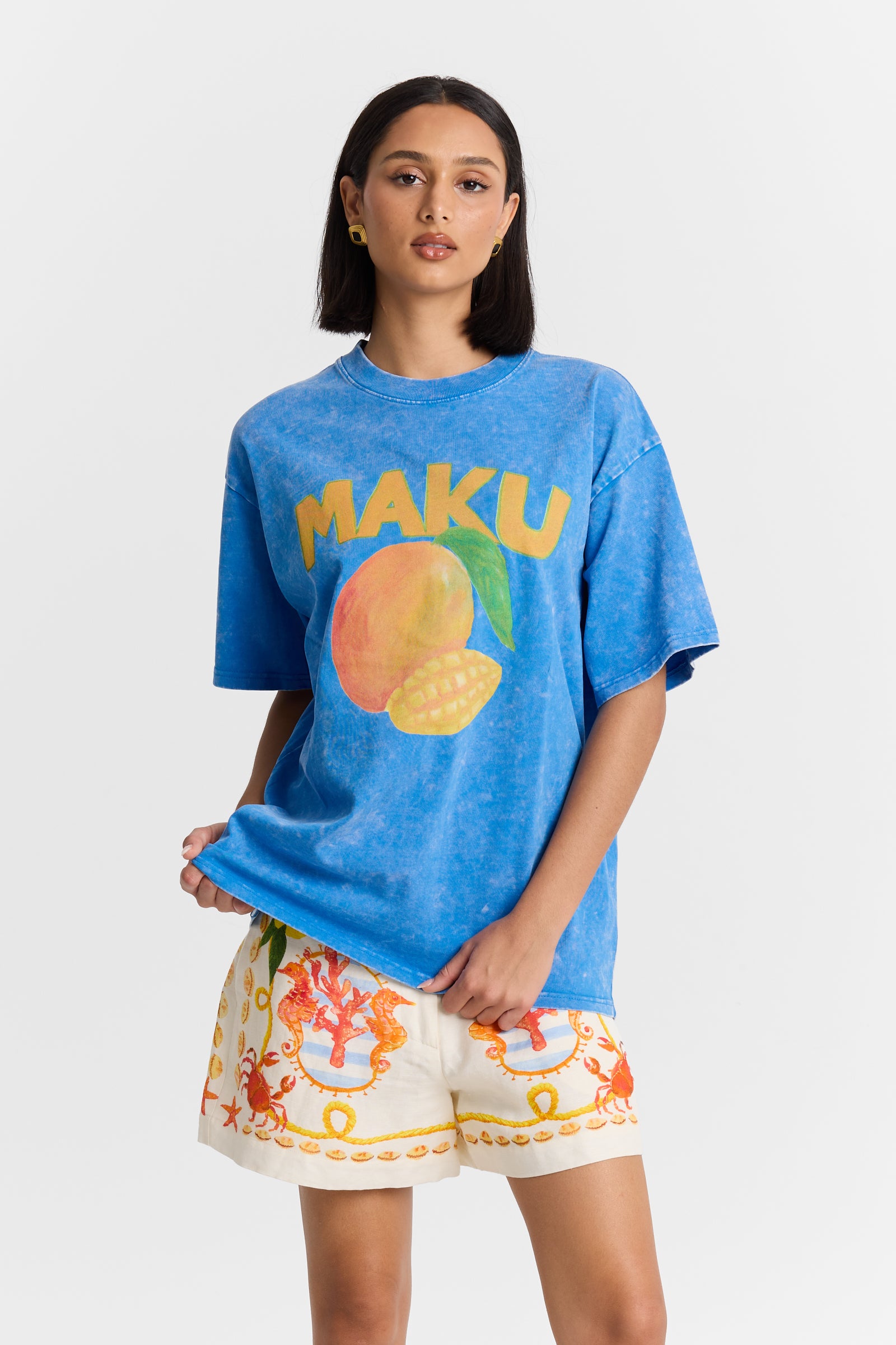 A Woman wearing a blue t-shirt with 'MAKU' and a mango graphic, paired with colorful shorts on a white background
