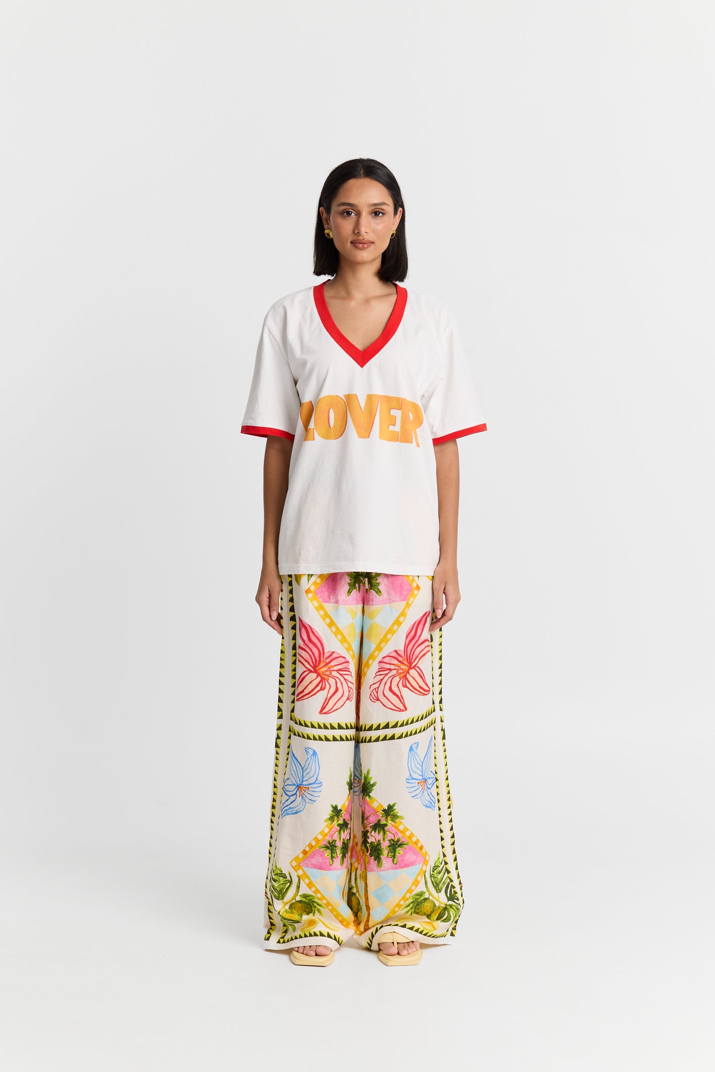Woman wearing a MAKU white VNeck shirt with 'LOVER' printed in orange and colorful pants on a white background
