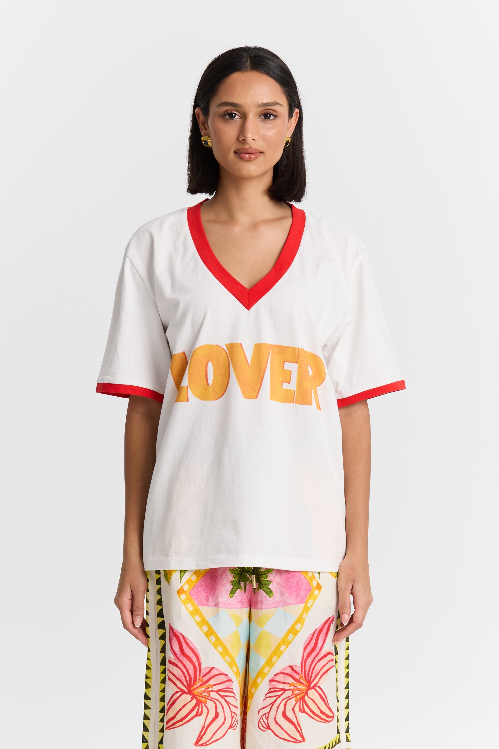 Woman wearing a MAKU white VNeck shirt with 'LOVER' printed in orange and colorful pants on a white background