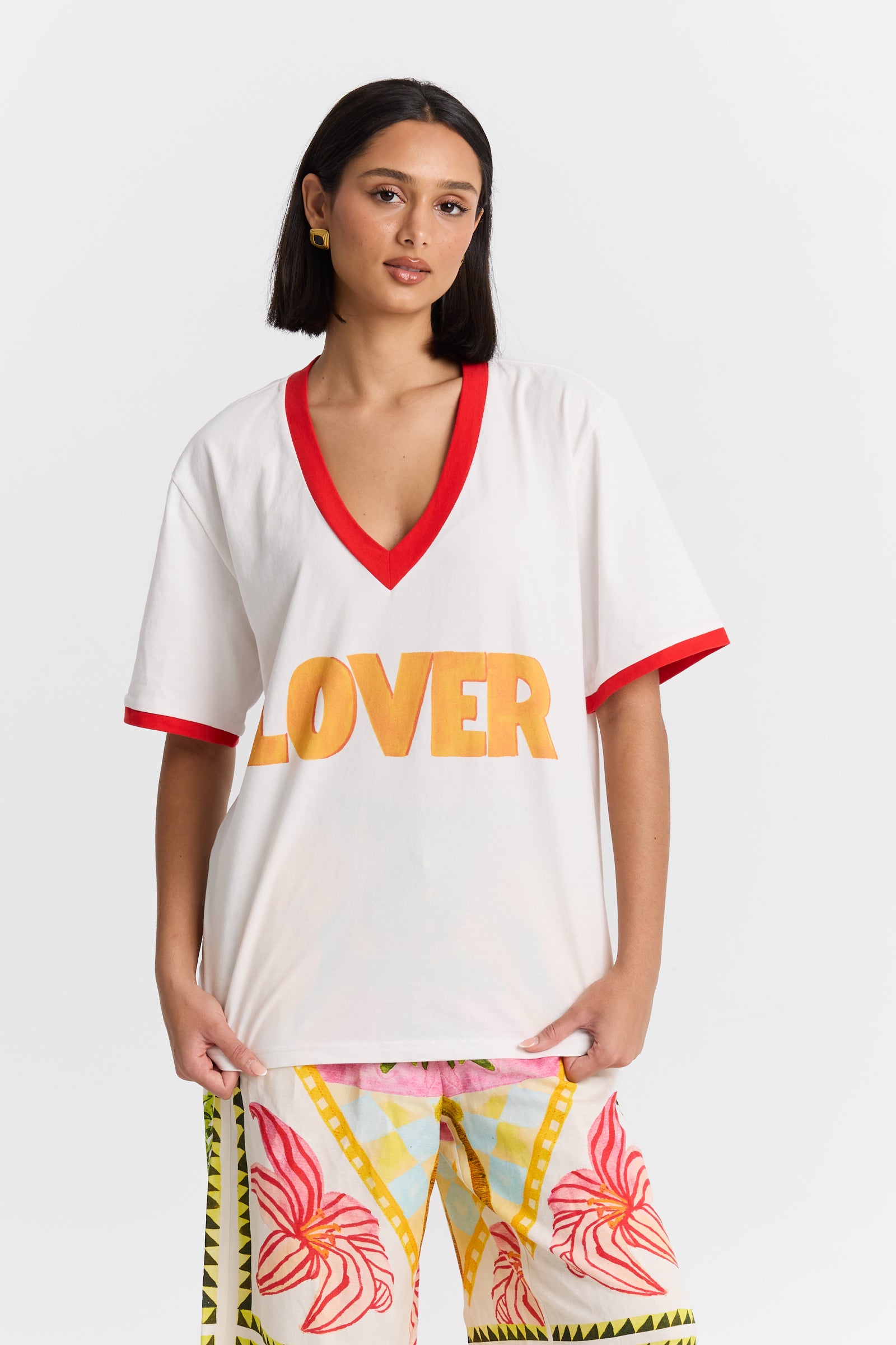Woman wearing a MAKU white VNeck shirt with 'LOVER' printed in orange and colorful pants on a white background