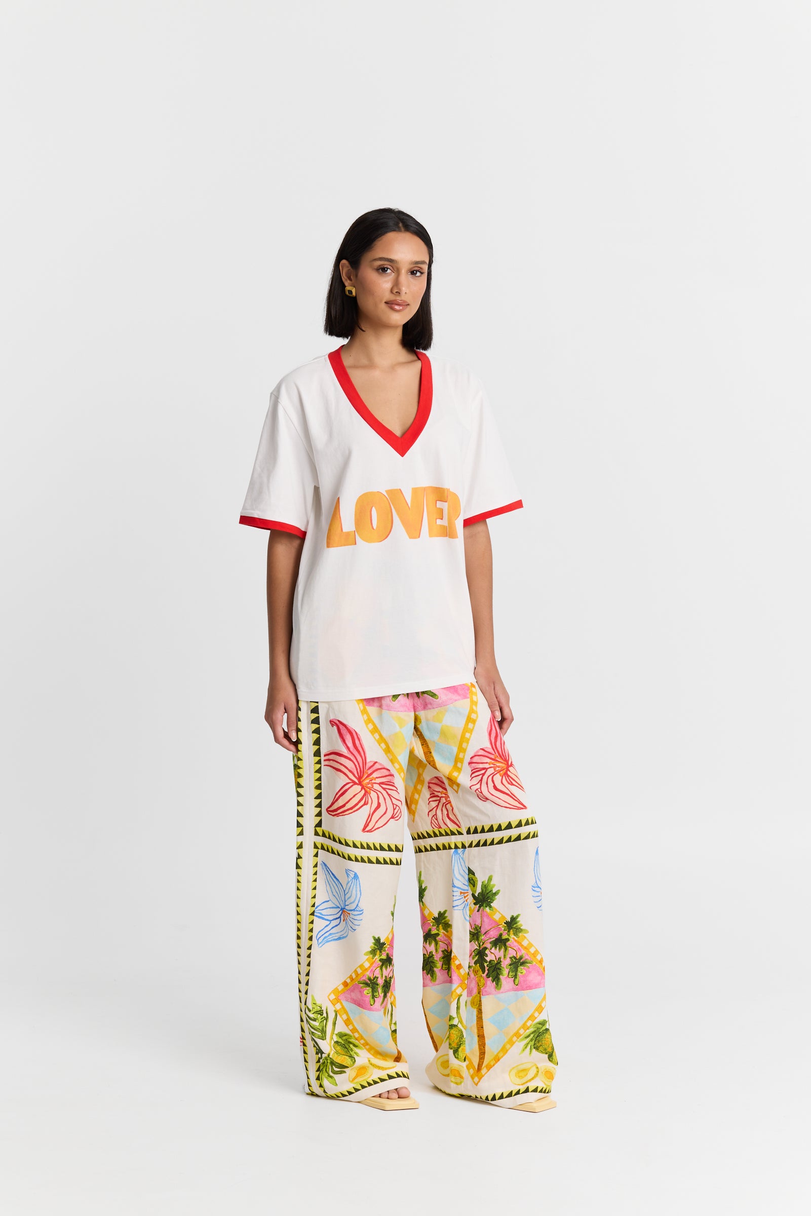 Woman wearing a MAKU white VNeck shirt with 'LOVER' printed in orange and colorful pants on a white background