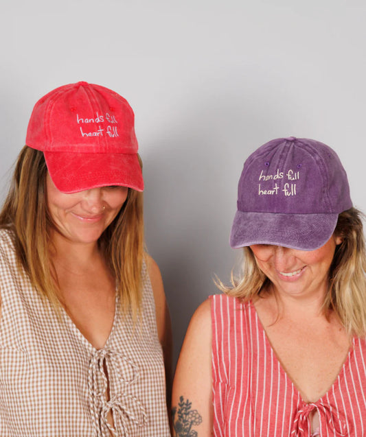 Two adults wearing matching baseball caps, one in red and the other in purple, with embroidered text on the front.
